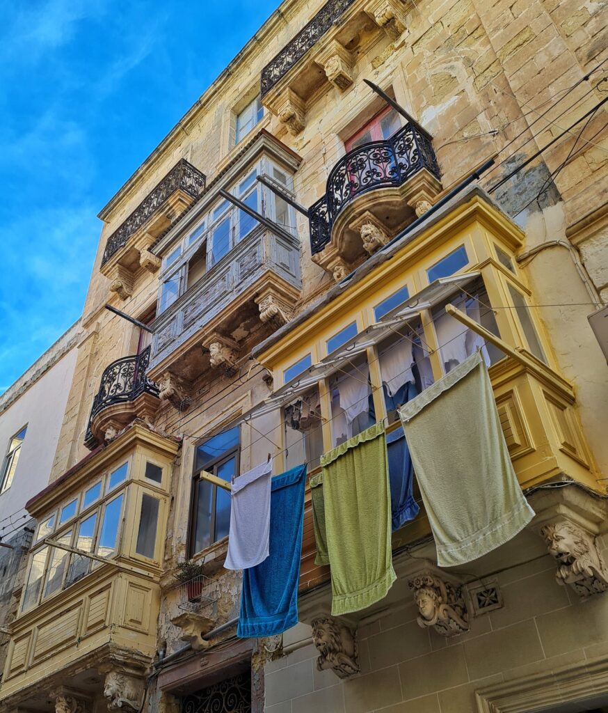 Ein Haus von unten schräg fotografiert. Es hat einige maltesische Balkone und einige mit schmiedeeisernen Gittern. Vor einem der unteren maltesischen Balkone hängen mehrere Handtücjer in verschiedenen Farben.