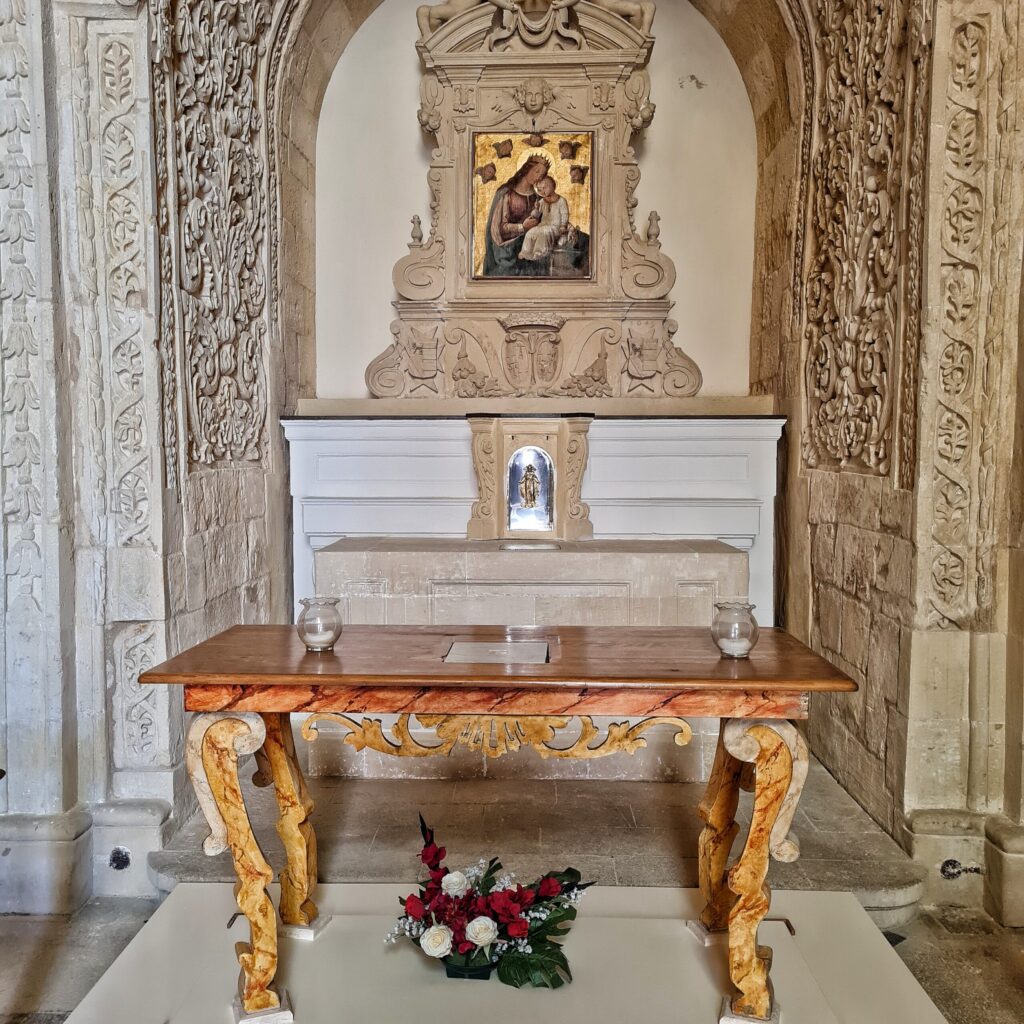 Ein schlichter Altar. Er besteht aus einem hellen Holztisch mit kunstvoll verzierten Beinen. Darunter liegt ein Blumengesteck. Auf dem Tisch zwei Kerzen in runden Gläsern. Im Hintergrund eine verzierte Nische. Dort hängt ein Gemälde von Maria mit dem Jesuskind.