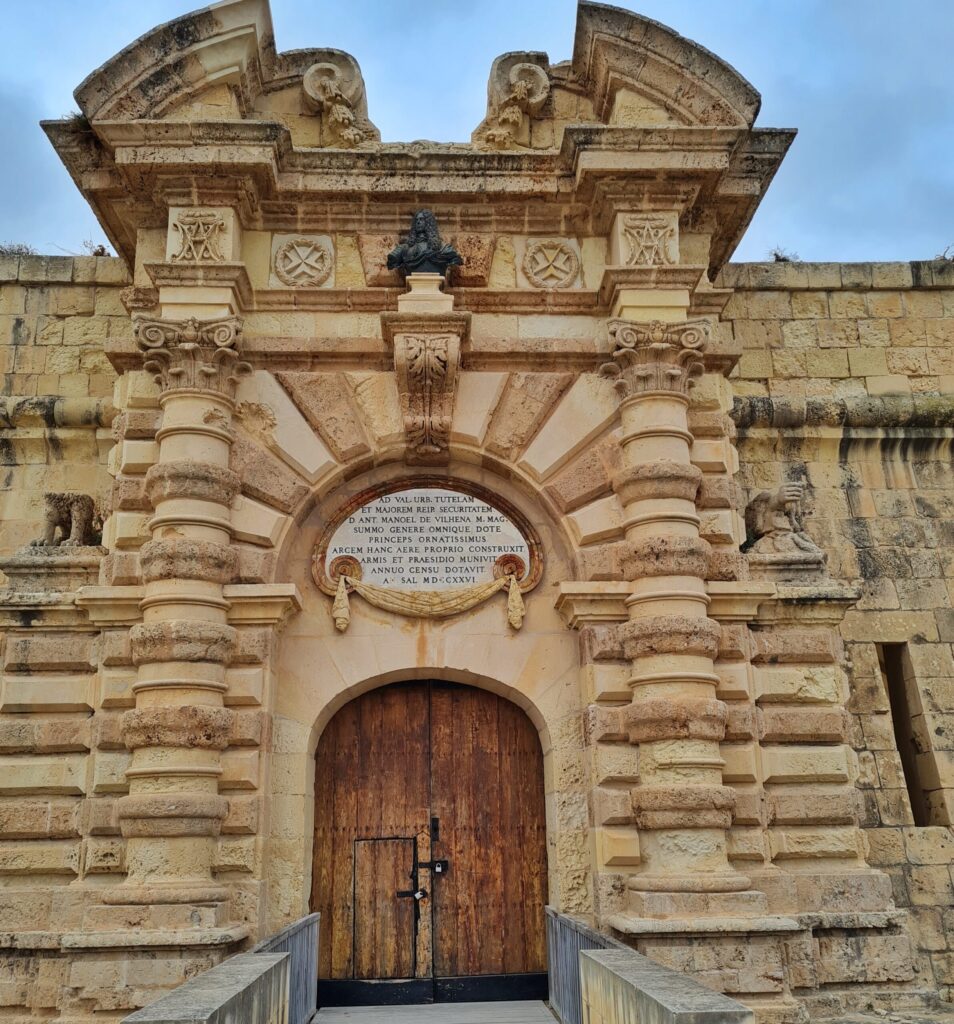 Ein hohes Tor aus hellem Stein mit vielen Verzierungen. Unten ein geschlossenes Holztor. Darüber eine runde große Plakette auf der etwas geschrieben ist (nicht lesbar). Oben die dunkle Büste eines Mannes mit langen lockigen Haaren.