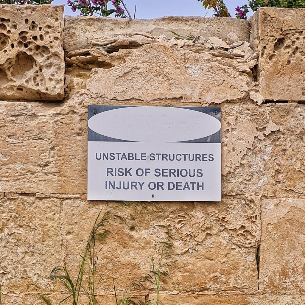Ein Teil einer beigen Steinmauer, teilweise porös. Darauf hängt ein Schild: "Unstable Structures. Risk of Serious Injury or Death".