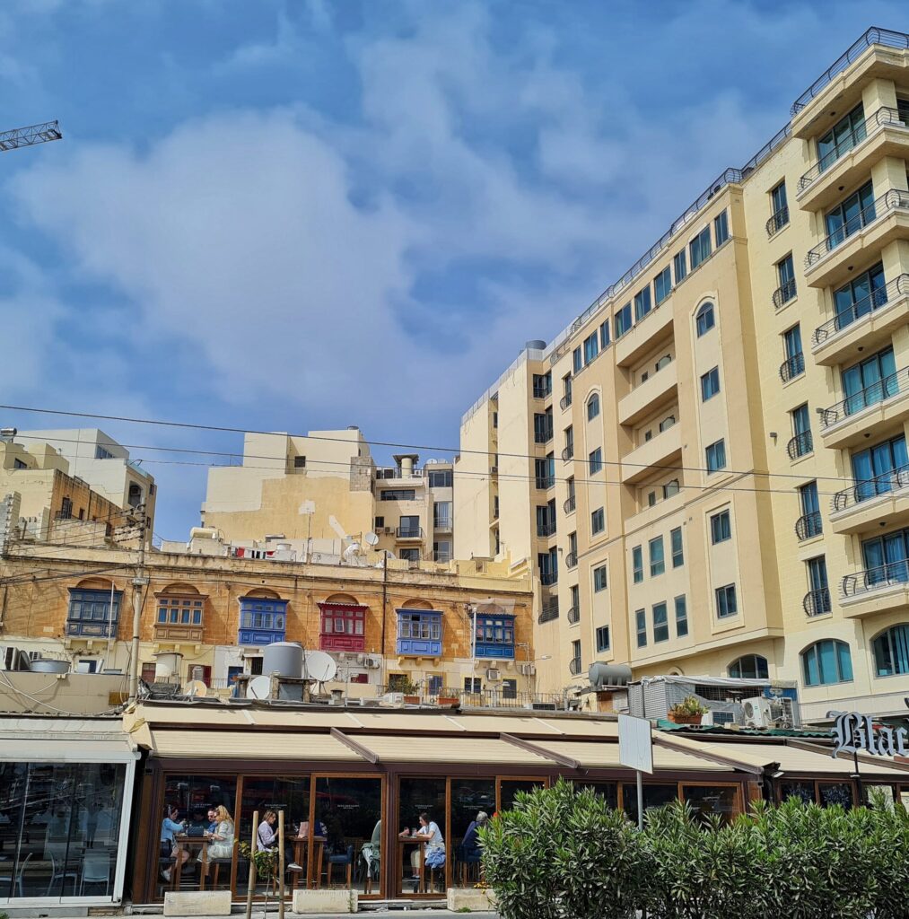 Rechts ein hohes modernes Gebäude mit vielen Fenstern und Balkonen, das quer steht. Im Hintergrund weitere hohe Gebäude. Im Vordergrund an einer Straße ein  flaches Gebäude mit einer Glasfront. Darin und davor Stühle und Tische, an denen Menschen sitzen. Dahinter ein längliches zweistöckiges Gebäude. Man sieht davon nur das obere Geschoß, an dem nebeneinader mehrere bunte maltesische Balkone hängen.