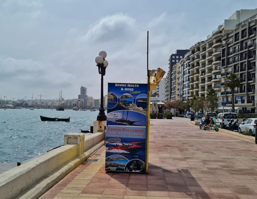 Eine breite Promenade. Links Wasser, das in der Sonne glitzert. Ganz im Hintergrund sind Schiffe und Gebäude zu sehen.  iRechts eine hohe Häuserreihe. Die Gebäude sind modern und haben viele Balkone. Zwischen den Häusern und der Promenade eine Straße mit Bäumen und parkenden Autos. Auf der Promenade steht ein großes Plakat, das mit Fotos für Schifffahrten wirbt.