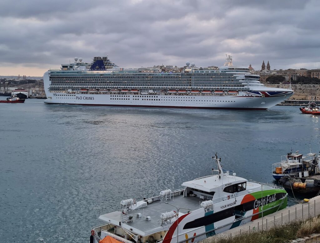 Ein weißes Kreuzfahrtschiff mit der Aufschrift "P & O Cruises" liegt am Kai vor Valletta. Vor dem Bug ein roter Schlepper. Auf der Seite des Hafens, von wo das Foto gemacht wurde, liegen eine Fähre mit den Aufschriften "Gozo Fast" und "Your Wisdom". Davor liegt ein anderes schwarzes Boot.