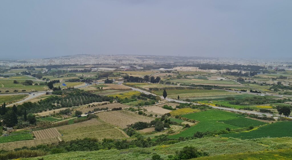 Ein weiter Blick in eine Landschaft. Im Vordergrund Felder, Straßen und Bäume. Im Hintergrund, etwas erhöht, eine Stadt, die jedoch nur undeutlich zu erkennen ist.