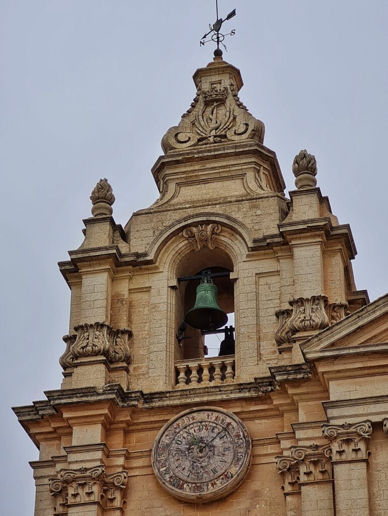 Einer der verzierten Kirchtürmchen mit der darin hängenden Glocke. Darunter ein Zifferblatt.
