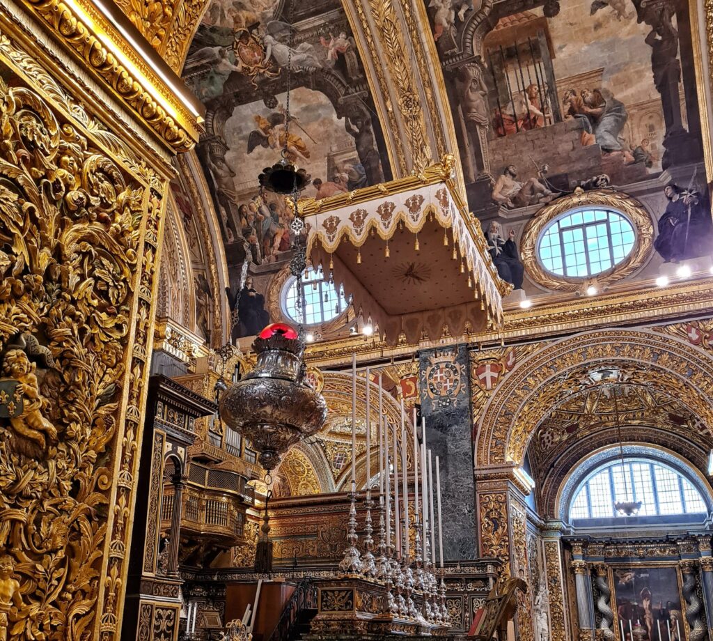 Blick auf einen Altar, über dem ein weiß-goldener Baldachin hängt. Davor stehen sehr hohe weiße Kerzen in  silbernen Leuchtern, und von der Decke hängt ein silbernes  großes Gefäß. Die Wände und Decken sind in Gold und mit Gemälden reich verziert.
