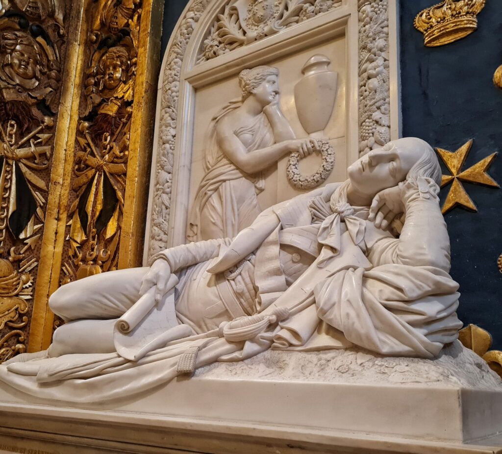 Eine weiße Statue eines seitlich  liegenden Mannes. Den einen Arm hat er aufgestützt, in der anderen Hand hält er ein leicht aufgerolltes Blatt Papier. Dahiner eine flache Darstellung einer Frau mit aufgestütztem Arm, die einen Kranz in der Hand hält. Darüber ein Gefäß mit Deckel.