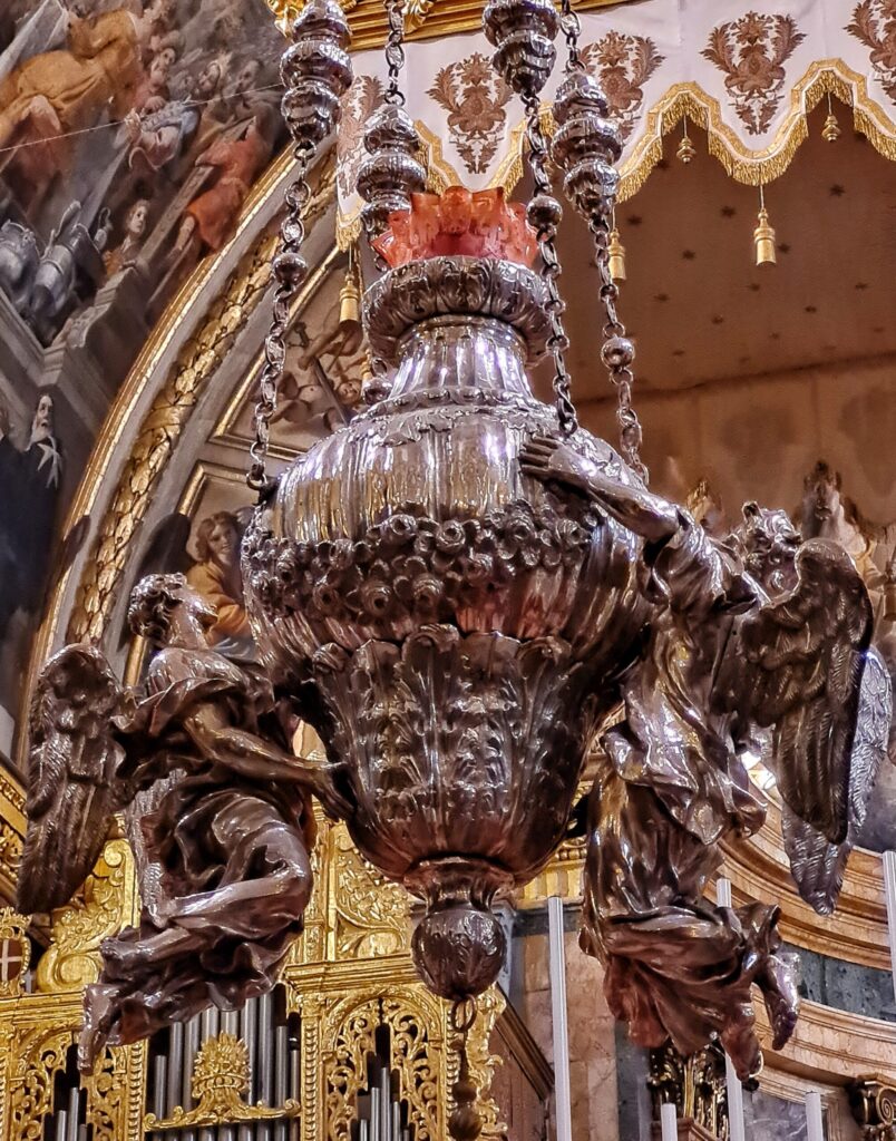Ein großesdunkel-silbernes Gefäß, das reich verziert ist. Oben sind verzierte Metallbänder zu sehen, an den das Gefäß offenbar hängt. Links und rechts an dem Gefäß hängen zwei kunstvoll gearbeitete fliegende 
Engel. Sie sind so dargestellt, als ob sie das Gefäß tragen würden.