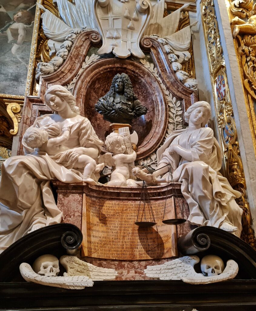 In einer runden Nische die dunkle Büste eines Mannes mit langem, gelockten Haar. Darum weiße Verzierungen. Davor drei weiße Statuen: links eine sitzende Frau, die ein Kind auf dem hält, rechts eine sitzende Frau, über deren Arm eine Waage hängt. In der Mitte ein kleiner sitzender Engel. Darunter links und rechts zwei Totenkopfdarstellungen.