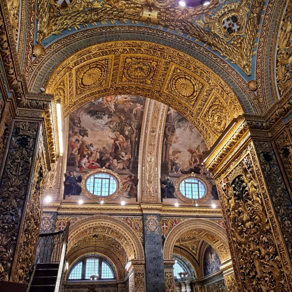 Blick in das Innere einer Kirche. Die hohen Rundbögen simd mit goldenen Mustern und Darstellungen reich verziert. Oberhalb der runden Fenster im oberen Bereich ein prächtiges Deckengemälde.