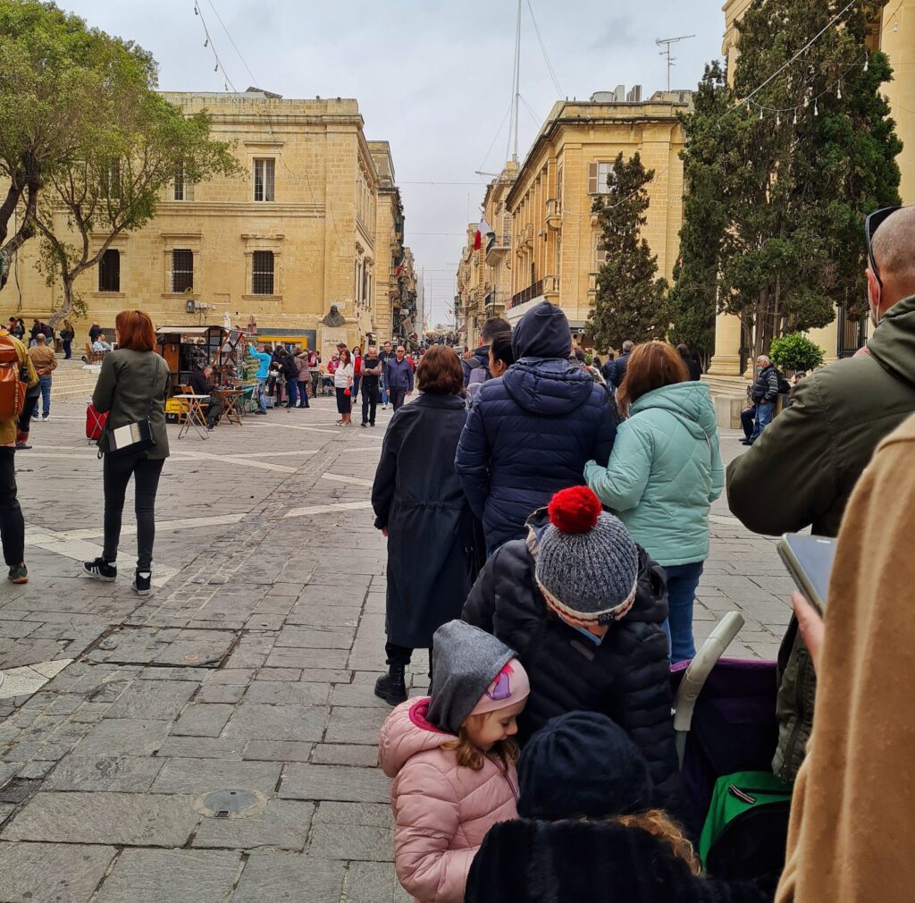 Eine belebte, schmale  Fußgängerzone. Links einige Straßenstände. Von hinten ist eine wartende Schlange von Menschen in warmen Jacken und mit Mützen fotografiert.