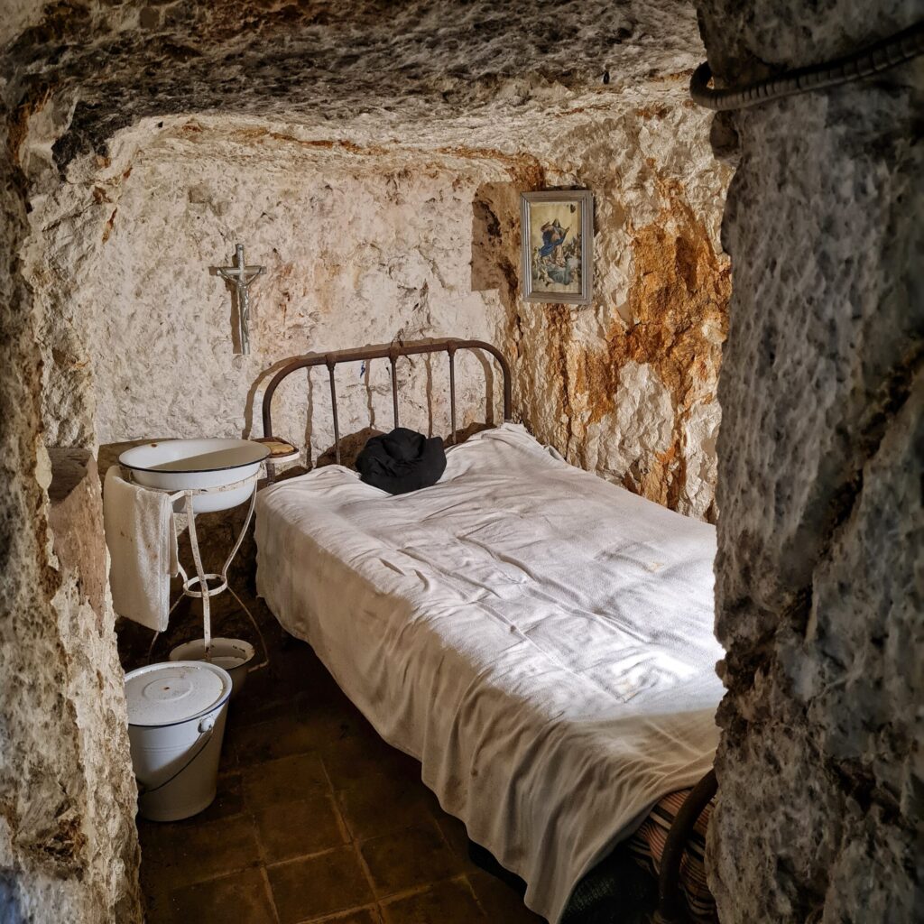 Ein kleiner offener Raum im Bunker. Darin ein einfaches Bett mit weißer Decke.Im Oberteil legt etwas Schwarzes. Daneben ein Gestell mit einer großen Emailleschüssel. Daran hängt ein weißes Handtuch. Daneben steht ein Emailleeimer mit Deckel. An der Wand ein Kruzifix und ein kleines Bild.