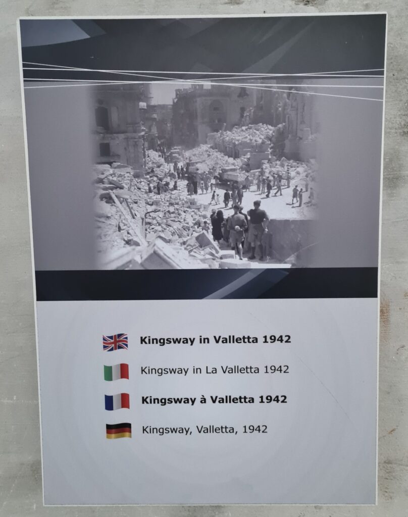 Ein schwarz-weißes Foto, das eine Straße mit zerstörten Gebäuden, Trümmern und Menschen zeigt. Darunter steht "Kingsway, Valletta, 1942".