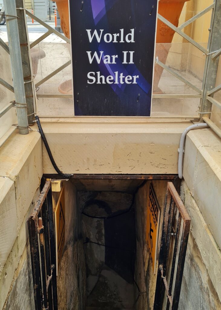 Blick nach schräg unten auf eine geöffnete Tür und einen Durchgang, der ins Dunkle führt. Darüber ein Schild "Wolrld War II Shelter".