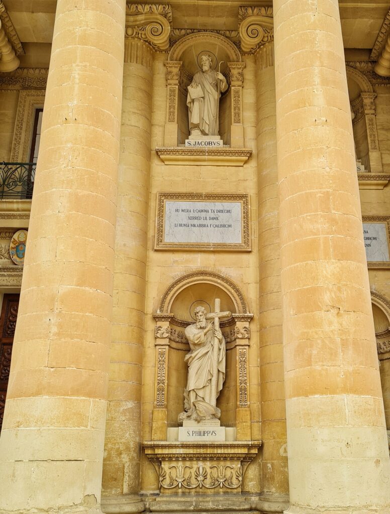 Hohe Säulen aus gelblichem Stein bilden eine Nische. In dieser sind zwei weitere Nischen, untereinander. In diser steht jeweils eine Figur in langen Gewändern und mit Heiligenschein. Die untere zeigt einen Mann, der ein Kreuz trägt, die obere einen Mann mit Bischofsstab. In der anderen Hand hält er ein Blatt Papier.