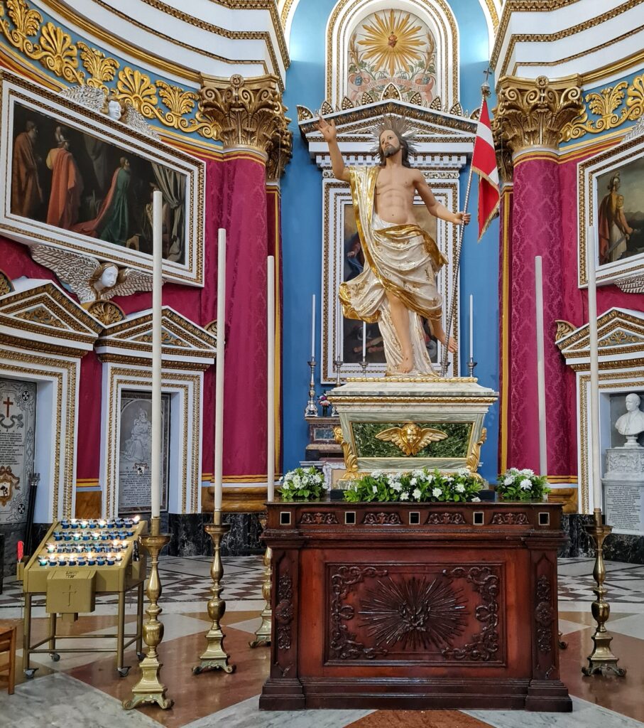 Auf einem Podest steht eine Christusstatue. Er ist nackt und hat um sich ein weißes Tuch mit goldenem Rand drapiert. Die rechte Hand hat er erhoben. In der linken Hand trägt er eine rot-weiße Flagge. Im Hintergrund Pfeiler und Wände mit lila Stoff und Gemälde. Im Vordergund eine Art Holzkasten mit Verzierungen. Daneben stehen mehrere sehr hoße weiße Kerzen in silbernen Leuchtern.