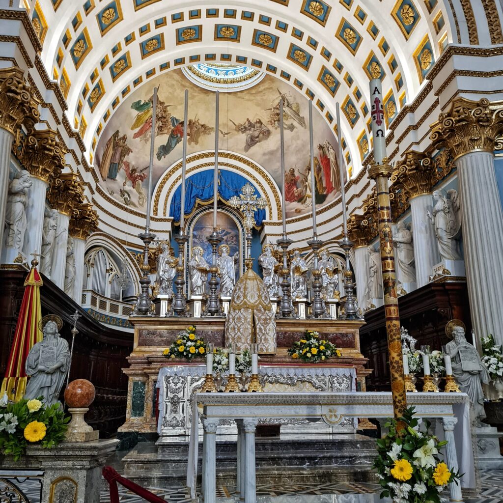 Ein Altar in weiß, kt gelben und weißen Blumengestecken geschmückt. Er ist umrahmt von weißen Pfeilern, oben mit Gold verziert, und weißen Statuen. Oben ist ein Bogen mit blau-goldene Mustern und ein Deckengemälde, das mehrere Menschen und Engel in farbigen Gewändern zeigt.