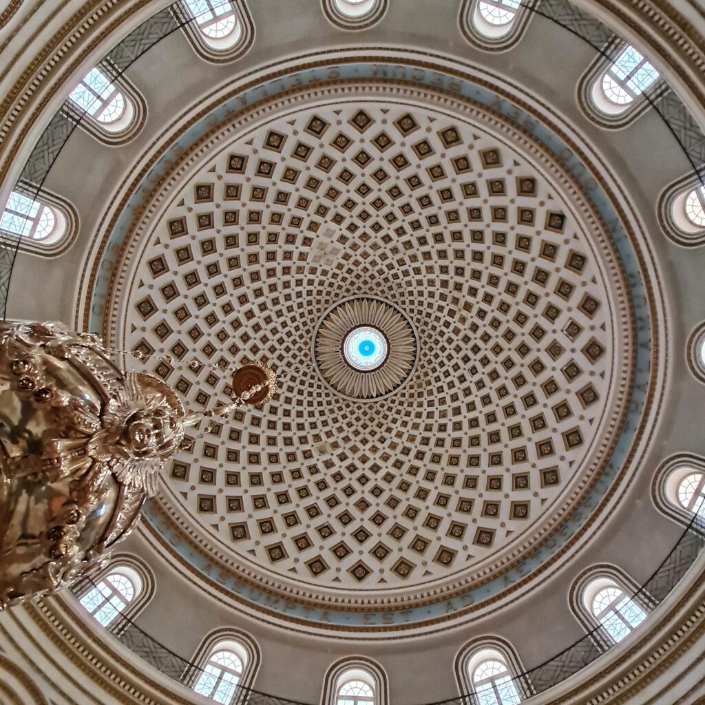 Blick auf die kreisrunde Kuppel von unten nach oben fotografiert. In der Mitte ist ein hellblauer Punkt. Darum herum bilden kleine Vierecke in Schwarz und Gold ein regelmäßiges Muster. Umrahmt wird dies von Fenstern. An der Kuppel hängt ein silbernes Gefäß mit Engelsköpfen, von dem auf dem Foto aber nur (im unteren linken Bereich) der obere Teil zu sehen ist.