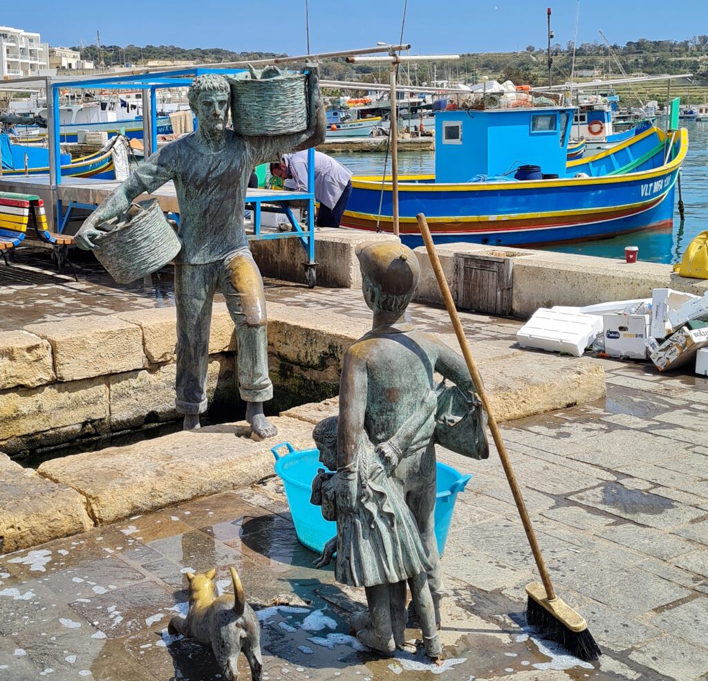 Auf einer Hafenpromenade, an der rechts viele blau-bunte Boote zu erkennen sind, zwei Figuren aus Metall. Die hintere zeigt einen Mann, der in Richtung auf die Kamera aus einer Öffnung im Boden steigt. Unter dem einen Arm und auf der anderen Schulter trägt er jeweils einen Korb mit Fischen. Vorne (von hinten zu sehen) ein Junge mit Mütze, der mit der linken Hand ein Mädchen festhält. Daneben eine Katze. Zwischen den Figuren steht eine blaue Plastikwanne, und am Jungen lehnt ein Besen.
