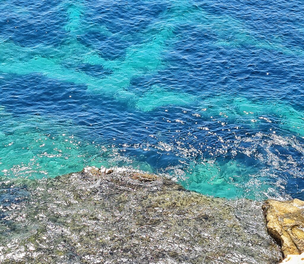Blick auf das Meer von oben. Unten im Foto flache Felsen. Darüber Meer in dunkel- und hellblau.