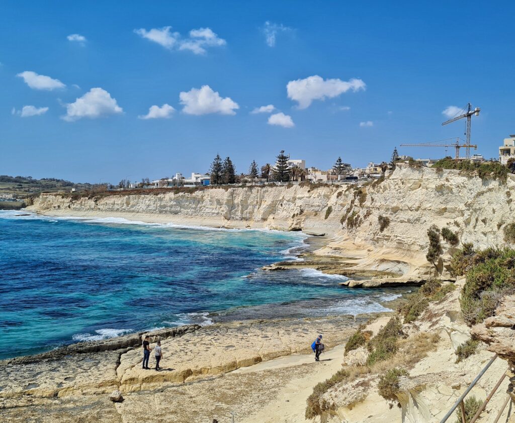 Eine Bucht gebildet aus weißen Felsen. Auf einnem flachen Uferteil stehen beziehungsweise laufen drei Menschen. Das Meer ist blau. Auf der oberen Kante der Felsen sind im Hintergrund einige Gebäude und Bäume und ein Kran zu erkennen.
