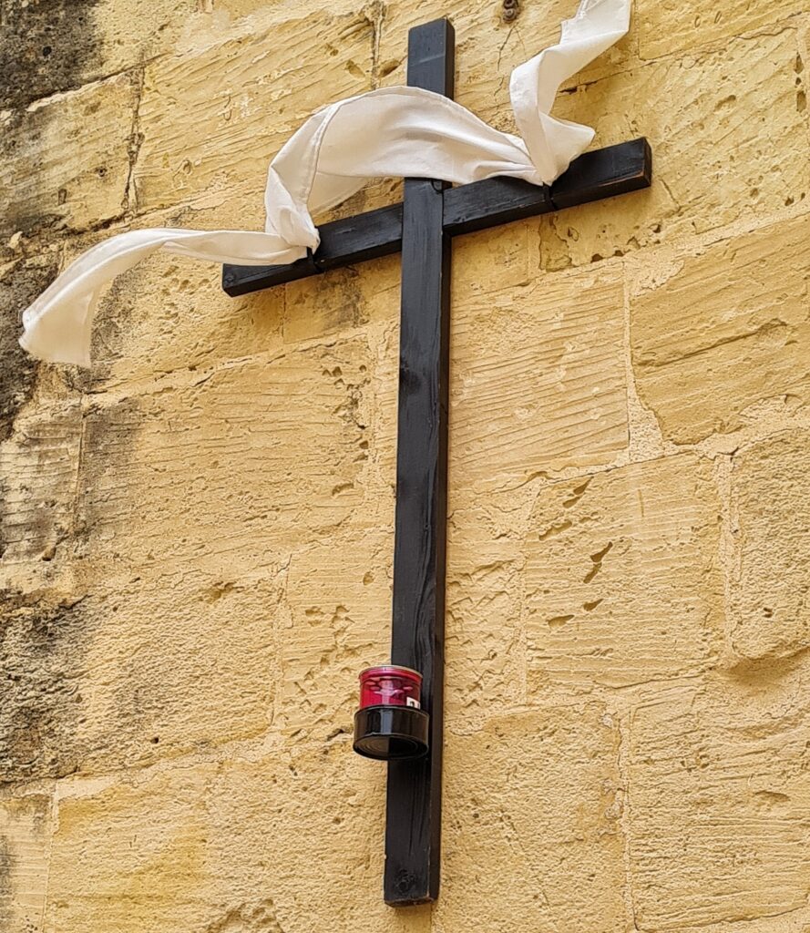 An einer gelblichen Hauswand ein großes Kreuz aus dunklem Holz. Daran ist oben ein weißer Flor angebracht und unten ein (nicht brennendes) rotes Grablicht.