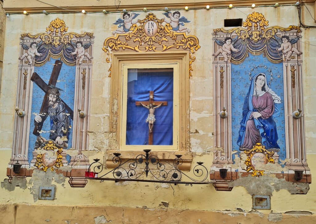 Eine gelbliche Hauswand. Links und rechts jeweils gemalte Darstellungen. Links ein bärtiger Mann, der ein Kreuz trägt. Rechts eine sitzende Frau mit Heiligenschein. Sie trägt ein rotes Kleid, einen blauen Umhang und ein weißes Tuch über dem Kopf. Sie hat die Hände gefaltet. In der Mitte in einer viereckigen, verglasten Nische eine Jesus-Darstellung am Kreuz. Um alle drei Darstellungen herum sind Rahmen mit Verzierungen und Engeln gemalt.