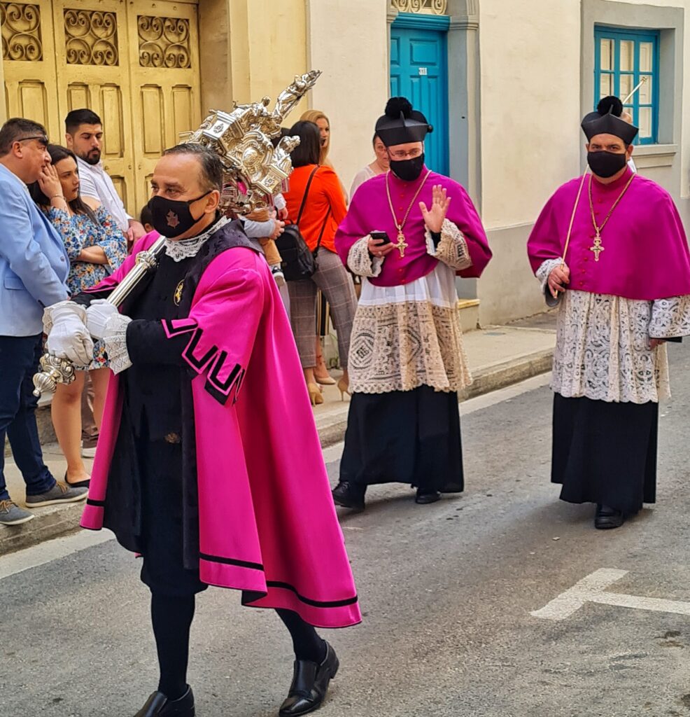 Auf der Gasse mit den zuschauenden Menschen laufen drei Männer. Der vordere trägt über schwarzer Kleidung einen dunkelpinken kurzen Umhang. Auf der Schulter trägt er eine silberne Stange mit etwas großem Silbernen daran. Hinter ihm laufen zwei Priester mit komisch aussehenden schwarzen Kopfbedeckungen. Sie tragen lange schwarze Priestergewänder, darüber ein helles Spitzengewand und darüber ein dunkelpinkes Oberteil. Um den Hals tragen sie goldene Kreuze. Einer hat ein Smartphone in der Hand. Alle drei Männer tragen schwarze Mund-Nase-Masken.