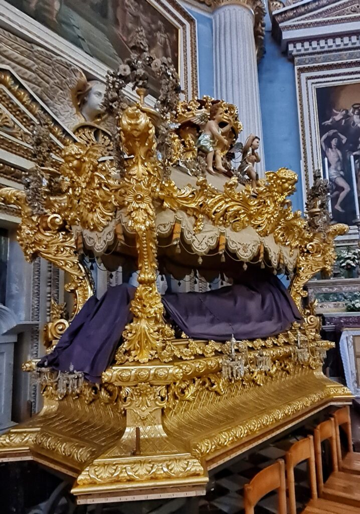 In einem Raum mit prächtigen Gemälden, Engelsfiguren, Säulen und goldenen Verzierungen an den Wänden steht etwas, das aussieht wie ein geöffneter goldener Sarg, wobei zwischen dem unteren Teil und dem Sargdeckel an den Ecken kleine Pfosten angebracht sind. Dazwischen liegt eine Figur, die vollständig von einem auberginefarbigen Tuch bedeckt ist.
