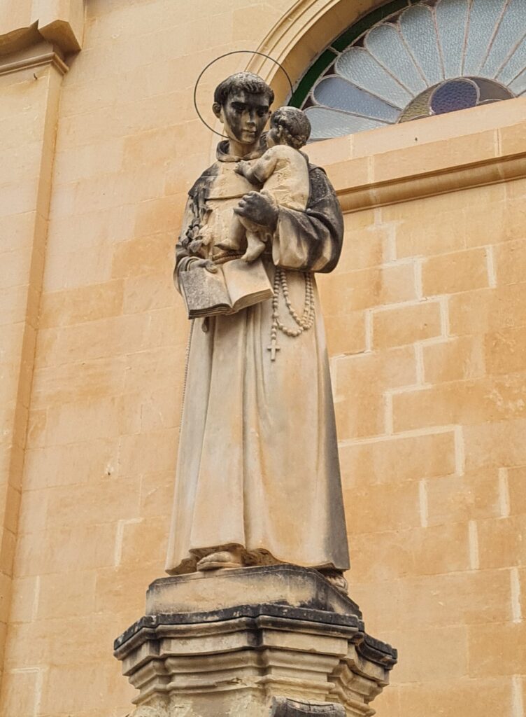 Eine gelbliche Gebäudewand mit einem halbrunden Fenster. Die Glasverzierung bildet eine Art Fächer. Davor steht die Statue eines jungen Mannes in Mönchskutte mit einem Heiligenschein. Ein Rosenkranz mit Kreuz hängt seitlich herab. Auf seinem linken Arm trägt er ein kleines Kind. Er und das Kind schauen sich an. In seiner rechten Hand hält er ein aufgeschlagenes Buch.