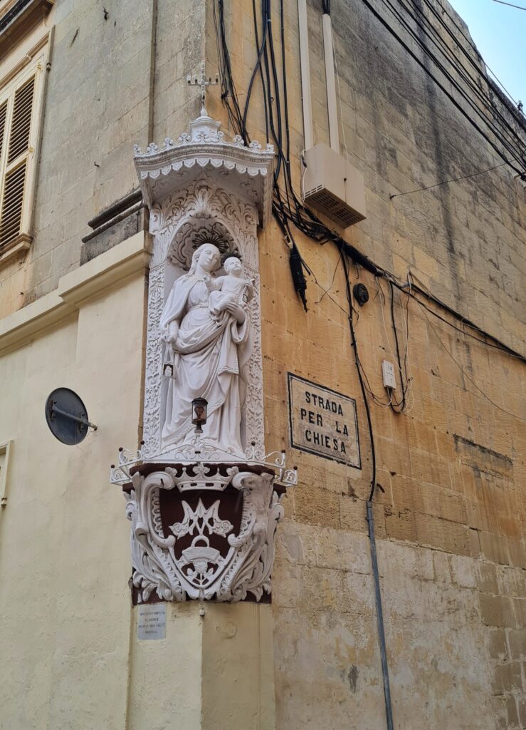 Die Hausecke eines gelblichen Gebäudes. Auf der rechten Wand ein kleines rechteckiges Straßenschild. In schwarzer Schrift auf weißem Grund steht dort "Strada Per La Chiesa". Direkt auf der Hausecke steht in einer Nische eine weiße Mariendarstellung mit Jesus auf dem Arm. Darunter Verzierungen, darüber ein kleiner Baldachin mit einem Kreuz darauf.