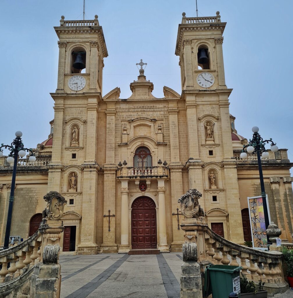 Die Kirche mit den beiden viereckigen Türmen, diesmal aus einer größeren Entfernung direkt von vorne fotografiert. Diesmal ist auch der Mittelteil der Kirche mit einem großen Holztor zu sehen. Darüber ein Balkon. Links und rechts ein geschwungener Zaun aus Stein und kugelförmige Straßenlaternen. Rechts ausserdem ein grüner Mülleimer.