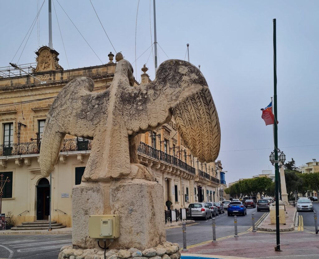 Eine Adlerstatue mit ausgebreiteten Flügeln aus Stein, von hinten fotografiert. Der Adler blickt auf eine Straße mit einigen Pkws, die durch eine längliche Verkehrsinsel getrennt ist. Im Hintergrund Bäume und Gebäude. Links ein großes zweistöckiges Gebäude aus gelblichem Stein. Vor den oberen Fenstern sind Balkone mit Metallgittern.