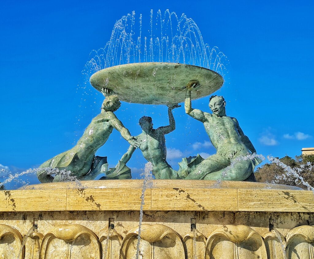 Der obere Teil eines Springbrunnens. Über dem äußeren Becken drei Figuren mit Fischflossen und männlichen nackten Oberkörpern. Seitlich spritzt Wasser auf sie. In jeweils einer Hand tragen sie gemeinsam eine große Schale, auf der Wasser im Bogen nach oben spritzt.