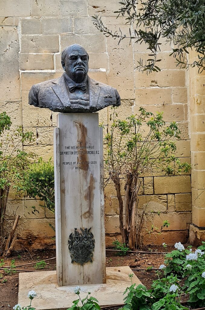 Eine Art Garten mit karger grüner Bepflanzung vor einer Steinmauer. Davor eine hohe weißliche Säule, auf der die Büste eines mißmutig schauenden alten Mannes steht. 