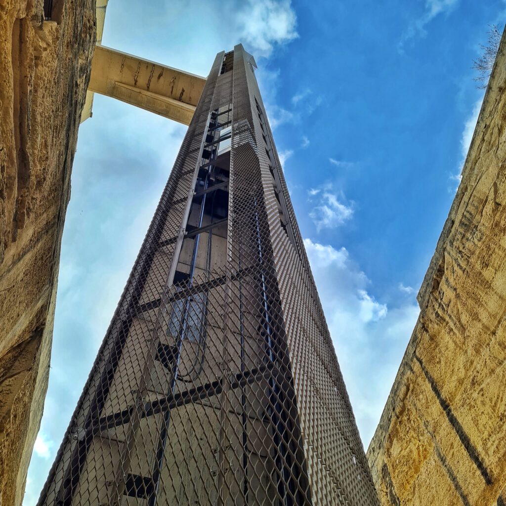 Der Fahrstuhl, von unten nach oben fotografiert. Er ist vergittert und man sieht eine  der Fahrstuhlkabinen.. Oben ist der Steg, der den Aufzug mit dem oberen Teil der Mauer verbindet, zu sehen.