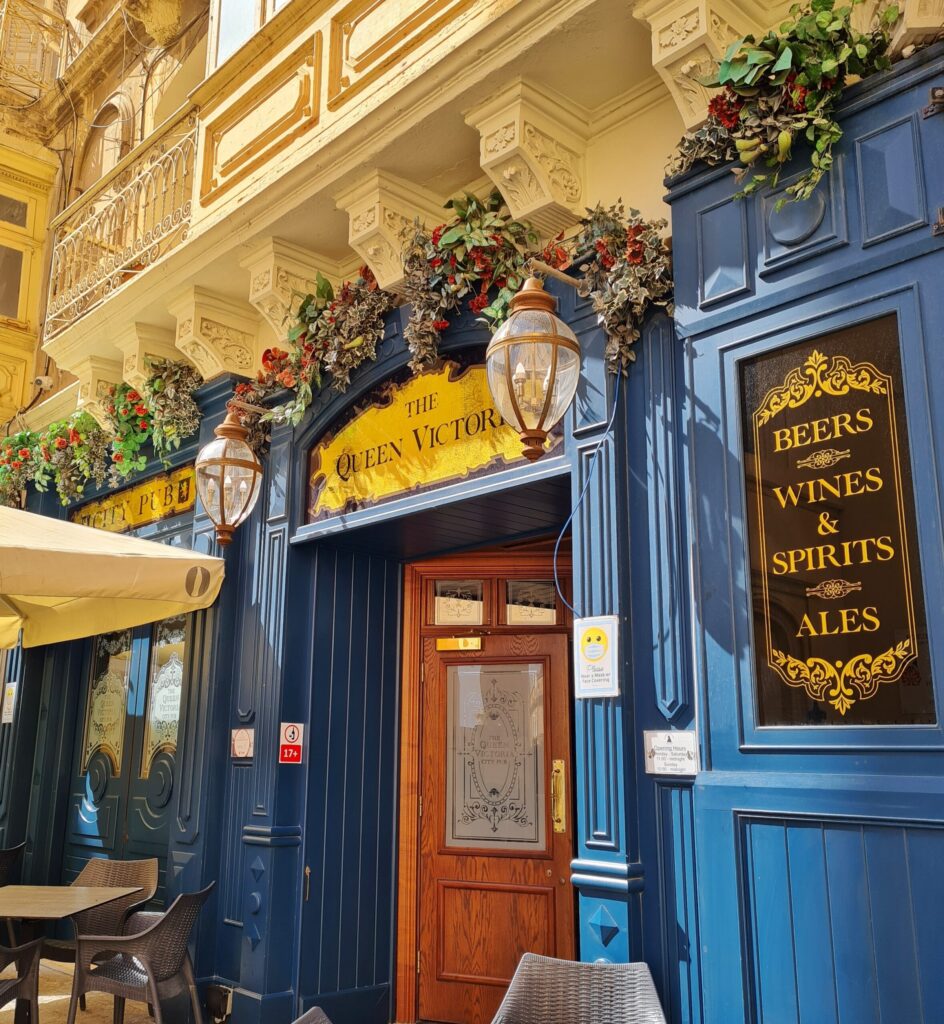 Der Eingang einer Gaststääte von außen fotografiert. Die untere Hauswand ist eine blaue Holzverkleidung. Über der braunen Eingangstür steht (in schwarz auf goldenem Hintergrund) "The Queen Victoria". Rechts ein Schild: "Beers Wines Spirits Ales" (in goldener Schrift auf schwarzem Hintergrund). Die blaue Holzverkleidung wird nach oben abgeschlossen durch künstliche Blumengestecke, die etwas herunterhängen. Darüber ist ein Balkon aus gelblichem Stein. Links unten sind ein Teil eines Sonnenschirmes und eines Tisches sowie zwei Stühle.