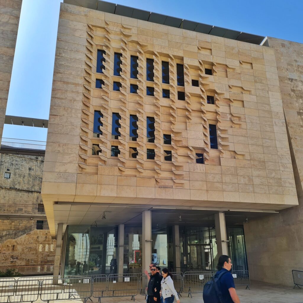 Ein quadratischer Gebäudeteil mit schmalen Fenstern, die ein Muster bilden. Er ruht auf schmalen Säulen; dahinter ist eine Glasfront.