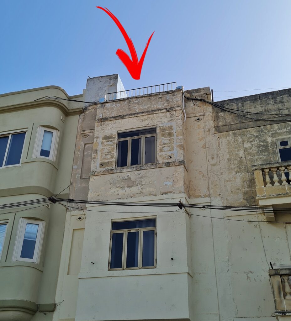 Der obere Teil eines schmales Hauses, das links und rechts an andere Häuser angrenzt. Zwei Fenster liegen übereinander. Oben ist das Dach flach und es ist ein Geländer daran. Ein großer roter Pfeil weist auf das Dach.