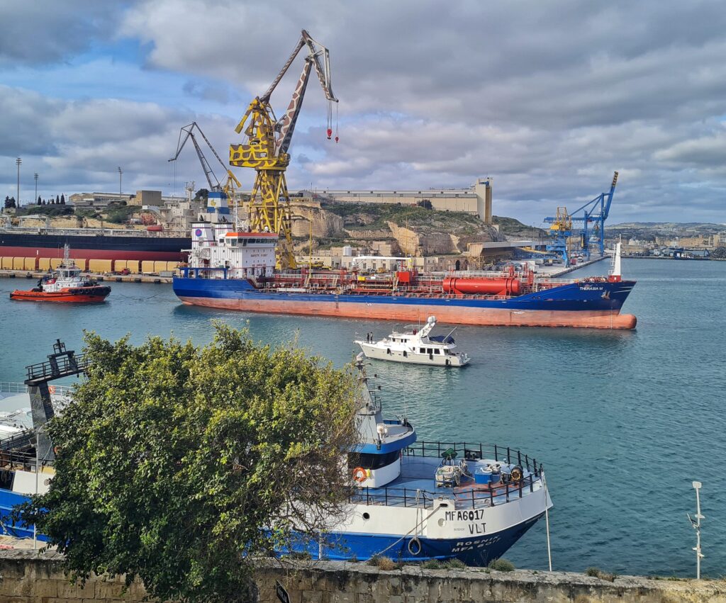 Vor dem Dock, in dem das große Schiff neben dem "Giraffen-Kran" liegt fährt ein großes Frachtschiff nach rechts, dahinter ein roter Schlepper. Nach links fährt eine kleine weiße Yacht. Am unteren Ufer liegt ein Schiff, größtenteils durch einen Baum verdeckt.