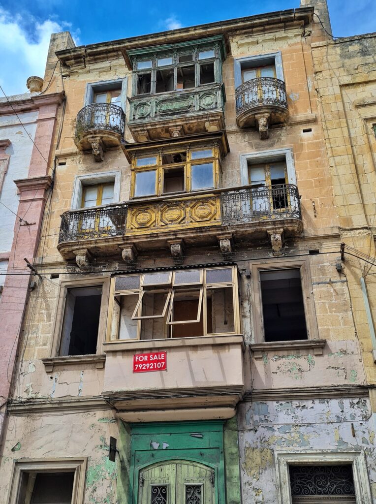 Ein ziemlich heruntergekommenes vierstöckiges Haus. In den beiden oberen Stockwerken maltesische Balkone. Vor den beiden Fenstern links und rechts davon jeweils klene Gitterbalkone. Darunter ein maltesischer Balkone, dessen Fenster leicht hochgeklappt sind. Die Fenster links und rechts davon haben keine Scheiben. Auf dem Balkon ein rotes Schild mit weißer Schrift: "For Sale" und eine Telefonnummer. Von der grünen Eingangstür und den unteren Fenstern ist nur der obere Teil zu sehen.