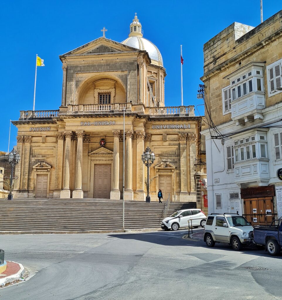 Eine beige Kirche an einer Straße. Rechts ein weiteres Gebäude und parkende Autos. Zur Kirche führen mehrere niedrige Treppenstufen empor. Die drei Türen (die mittlere mit Säulen davor) sind geschlossen. Darüber eine Dachterrasse mit einem Aufbau in der Mitte. Dort weht eine gelb-weiße Flagge. Im Hintergrund ein runder Turm mit einem kuppelförmigen Dach und einem Türmchen darauf.