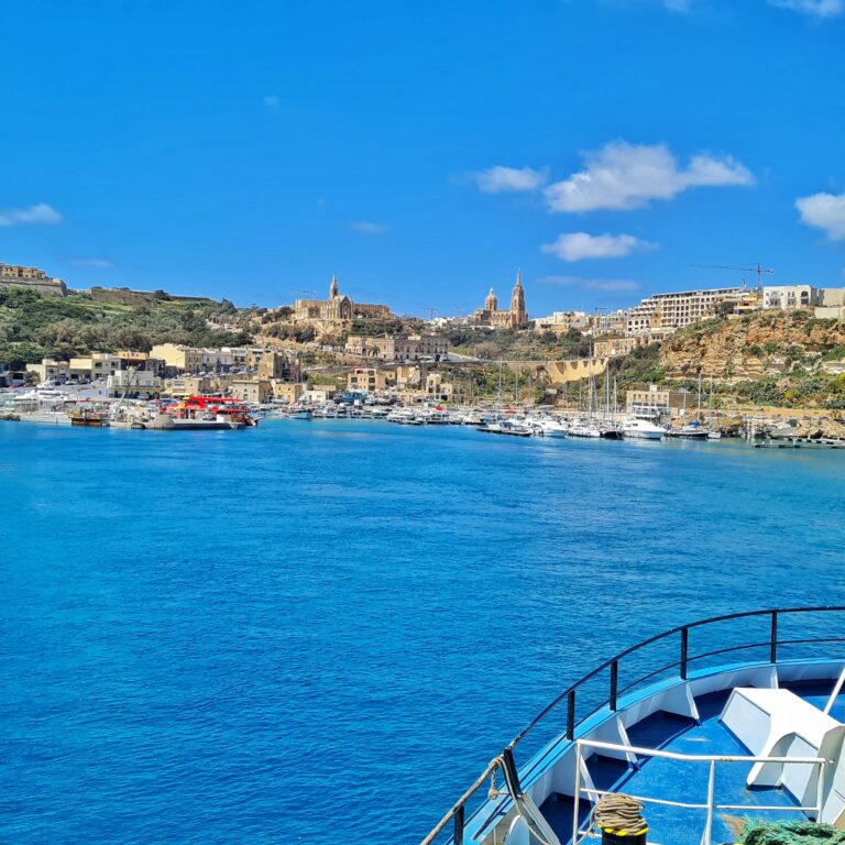 Blaues Wasser.Unten rechts ist ein Teil des Bugs eines großen Schiffes zu sehen. Das Deck ist hellblau. Im oberen Teil des Gebäudes eine Ortschaft, die sich auf die Felsen und Hügel erstreckt. Die meisten Häuser sind niedrig. Rechts oben ist aber ein modernes, langgestrecktes Gebäude zu sehen. Links davon zwei große Kirchen.