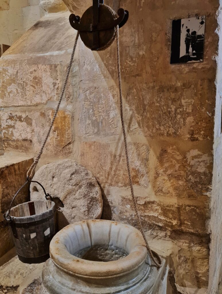 Eine helle Mauer aus Steinen im Inneren. Oben hängt eine große Seilwinde. Die beiden Seile führen nach unten zu einem Holzeimer und einem großen Steingefäß, das oben eine vasenartige Form hat. Es ist nach oben offen. An der Wand hinter dem Holzeimer lehnt eine runde Steinplatte. Oben rechts an der Wand ist ein Schwarz-Weiß-Foto, auf dem Menschen zu sehen sind.in