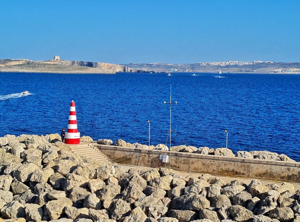 Im Vordergrund eine Kaimauer, davor sind zum Wasser hin, Steine verteilt. Der Weg hinter der Kaimauer führt über Stufen zu einem kegelförmigen Turm, der rot-weiß gestreift ist. Daneben steht eine Frau. Der Turm ist etwa dreimal so groß wie sie. Neben dem Weg sind weitere Steine aufgetürmt. Hinter der Kaimauer blaues Meer, auf dem einige weiße Schnellboote fahren. Links hinten ist eine Landzunge mit Felsen und einem kleinen viereckigen Turm. Ganz hinten ist ebenfalls Land zu sehen, wobei man auf einem Hügel Bebauung erkennen kann.