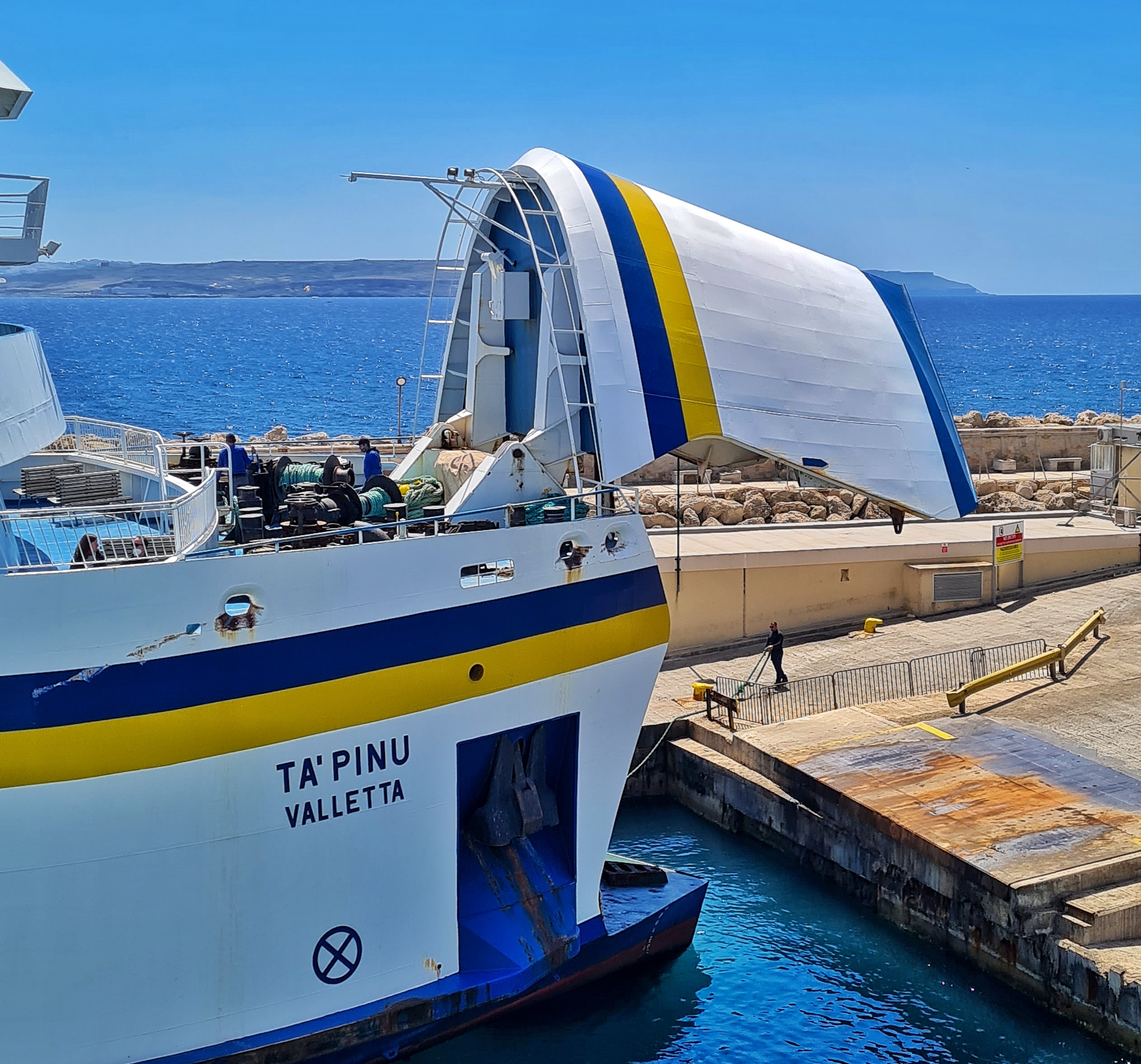 Ein großes Schiff (weiß mit einem blauen und einem gelben Streifen) fährt auf einen Hafenanleger in Form einer Rampe zu.Der Bugteil ist nach oben geöffnet. Auf dem Schiff steht TA'PINU VALLETTA. Hinter dem Schiff eine Hafenmole, dahinter blaues Meer und ganz im Hintergrund flaches Land.