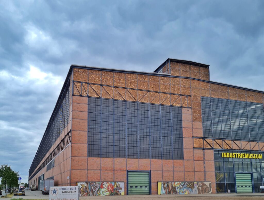 Eine riesige rötliche Fabrikhalle, etwas seitlich fotografiert, so dass man die Vorderseite und die linke Gebäudeseite sieht. Sie hat große Fensterflächen mit Gitter. Vorne links steht ein Schild mit der Aufschrift "Industriemuseum" Die gleiche Aufschrift steht in gelben Buchstaben über dem verglasten Eingangsbereich.