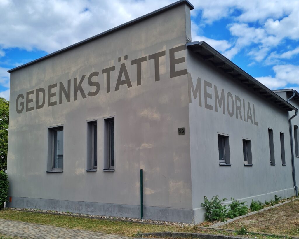 Ein hellgraues niedriges Gebäude mit einigen schmalen Fenstern. Auf der einen Seite steht in großen Buchstaben: Gedenkstätte, auf der anderen: Memorial.