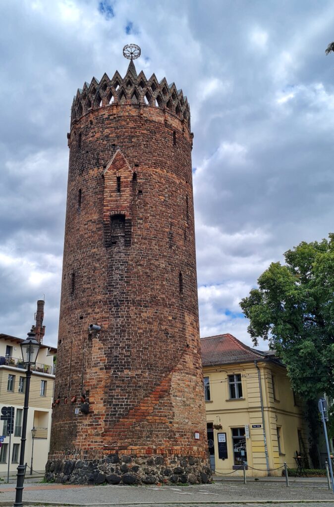 Ein hoher runder Backsteinturm vor einigen hellen Gebäuden mit Wohnungen und Läden. Er hat nur wenige schmale Öffnungen. Oben eine Umrandung aus zwei gezackten Linien. Darin ein radförmiger Gegenstand auf einer Spitze.