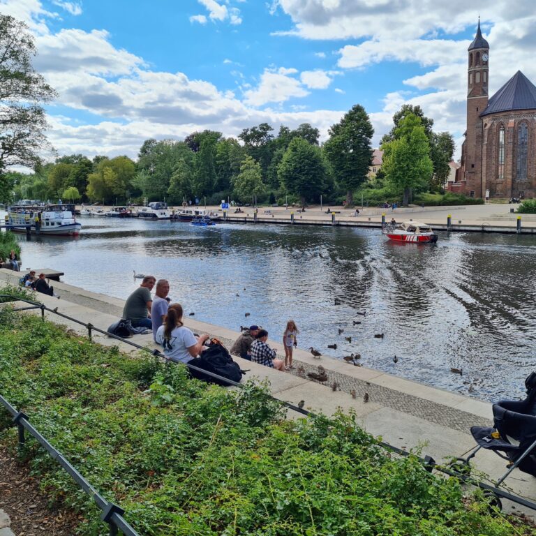 Ein Fluß, auf dem mehrere Schiffe und Boote fahren. Auf der anderen Seite eine Kirche. Daneben Bäume und davor eine breite Uferpromenade. Auf der Seite, von wo das Foto aufgenommen wurde, ein von einem niedrigen Metallzaun umgebenes Beet mt niedrigen Büschen. Davor führen breite Stufen zum Wasser hinab, auf denen Menschen sitzen. Unten am Wasser mehrere Enten und ein stehendes Kind. Rechts ist ein Teil eines Kinderwagens zu sehen.